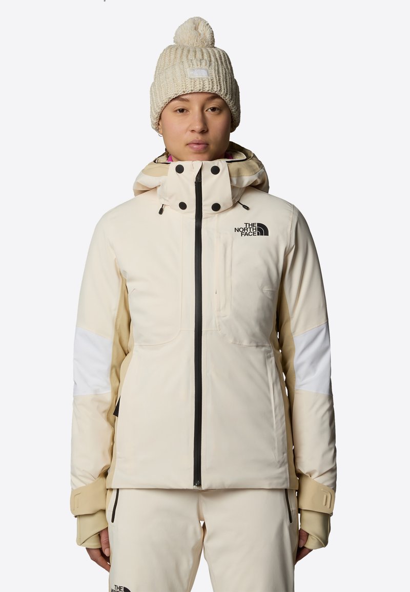 Giacca imbottita beige con zip nera e accenti delle tasche, con pannelli bianchi sulle braccia e colletto alto. Berretto lavorato a maglia color crema con pom-pom.