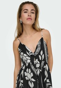 Zwarte jurk met witte bloesem- en bladerenpatronen, V-hals, verstelbare dunne bandjes en een geplooide taille. Model draagt bloemenoorbellen.