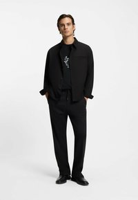 Chemise noire boutonnée portée sur un t-shirt graphique noir, associée à un pantalon noir à fines rayures et des chaussures noires sans lacets. Le mannequin se tient dans un cadre neutre.