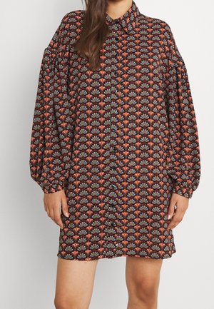 Kobieta ubrana w mini sukienkę na guziki z długim rękawem, z kolorowym, powtarzającym się kwiatowym wzorem w ciemnych tonach.