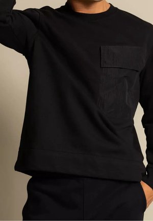 Mann trägt ein schwarzes Langarmshirt mit einer großen, strukturierten Brusttasche, eine Hand erhoben und die andere in der Hosentasche, beiger Hintergrund.