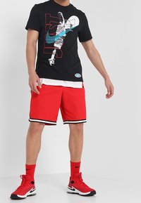 Černé tričko s grafikou basketbalu a červeným nápisem, doplněné červenými šortkami s černými a bílými pruhy a černými sportovními botami.