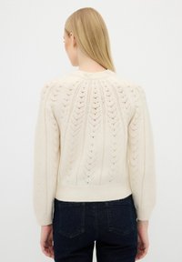 Maglione lavorato a maglia color crema con un design a traforo decorativo, polsini e orlo a coste. Presenta dettagli decorativi con perle nella parte posteriore del collo.