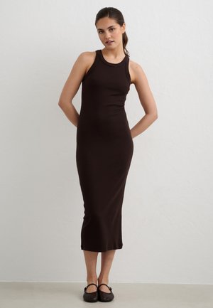 Frau mit dunklem Pferdeschwanz, die ein ärmelloses schwarzes Midikleid und schwarze Ballerinas trägt, steht vor einer schlichten hellen Wand.