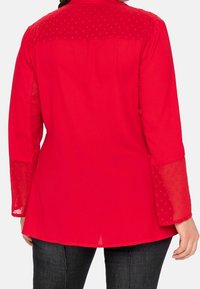 Blouse rouge avec manches longues transparentes ornées d'un motif à pois et dos uni. Ourlet légèrement plus long. Tissu lisse avec une coupe décontractée.