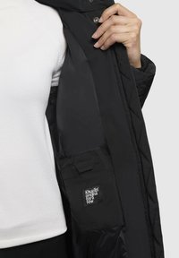 Cappotto trapuntato nero con chiusura a bottoni a pressione. Presenta una tasca interna con etichetta del logo. La texture visibile è liscia e leggermente lucida.