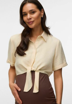 Naine, kellel on pikad tumedad juuksed, seljas lühikeste varrukatega kreemjas särk, mis on taljest seotud, ja kõrge vöökohaga pruun seelik, puudutades ühe käega puusa.