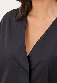 Černý oděv s v-hloubkou, jemné textury, s mírným záhybem a minimálně viditelným švem; zahrnuje malou náprsní kapsu.