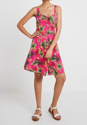 Frau in ärmellosem, knieumspielendem, rosa Blumenkleid mit Rüschendetail und weißen Plateausandalen, stehend mit der Hand in der Hüfte.