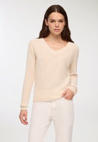 Beżowy sweter z dzianiny z dekoltem w kształcie V, ściągaczami przy mankietach i u dołu. Lekka faktura, lekko luźny fason, noszony z jasnymi dżinsami.