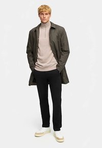 Olivgrön lättviktsjacka över en beige tröja med dragkedja i halsen, kombinerad med svarta byxor och vita sneakers med beige detaljer.