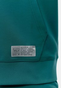 Tyrkysová mikina s kapucňou vyrobená z pleteného materiálu, s neoprenov�ým efektom, štyri smerový streč, elastická textura a antibakteriálnym označením.