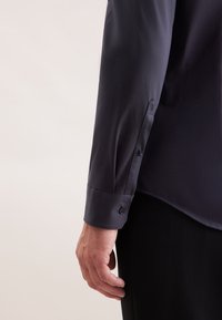 Camicia a maniche lunghe blu navy scuro con una texture liscia, polsini con bottoni e orlo curvo. Mano appoggiata su una superficie nera testurizzata.