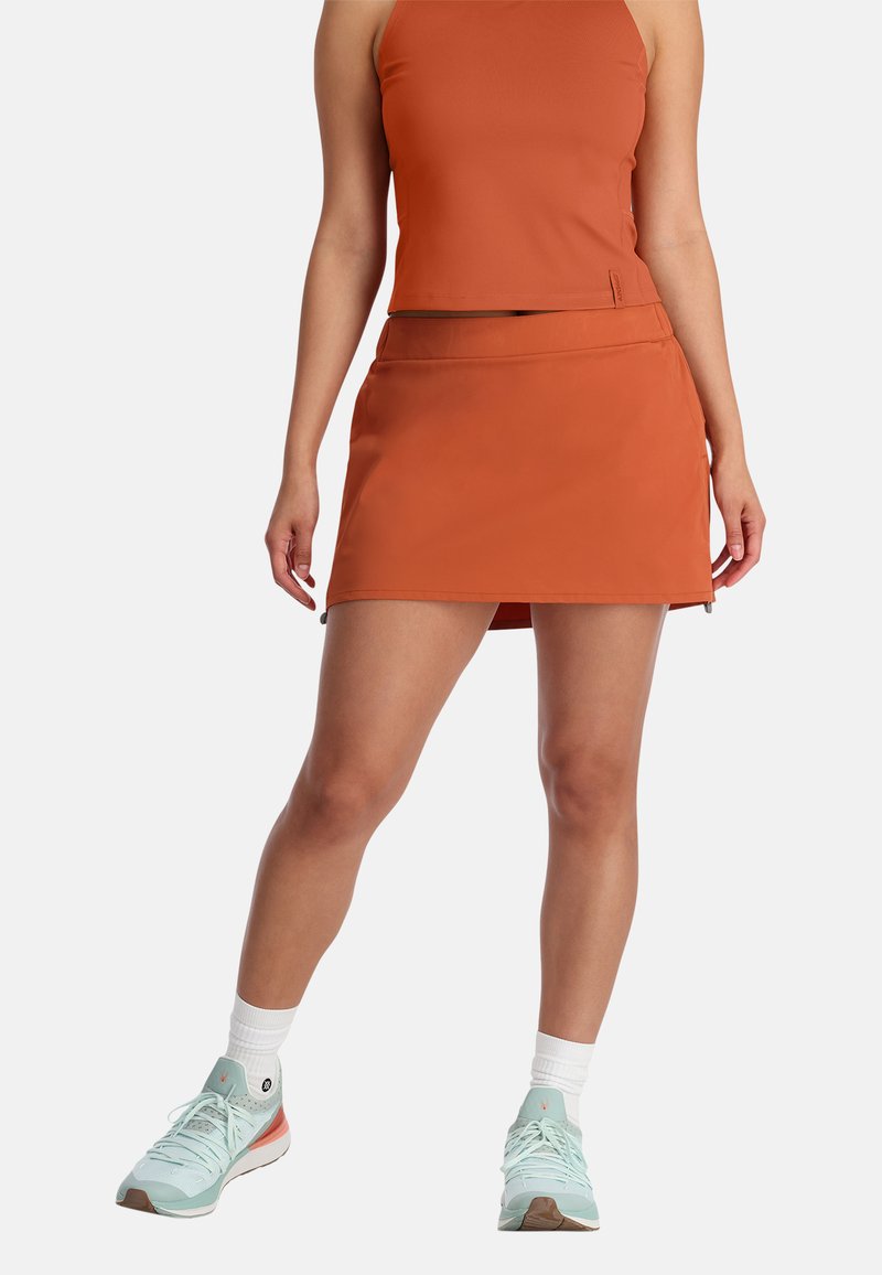 Oranje, aansluitende sportrok met een gladde textuur en een lichte glans; gecombineerd met een bijpassende crop top en mintgroene sneakers.