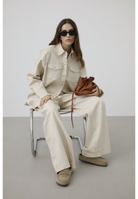 Sieviete valkā bēša krāsas oversized krekli un bikses, bēšas apavus, melnas saulesbrilles, sēž uz metāla krēsla, turot brūnu ādas somiņu.