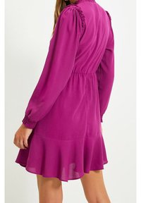 Robe fuchsia avec un col et des manches à volants, taille cintrée et ourlet évasé, fabriquée en tissu léger à texture lisse.