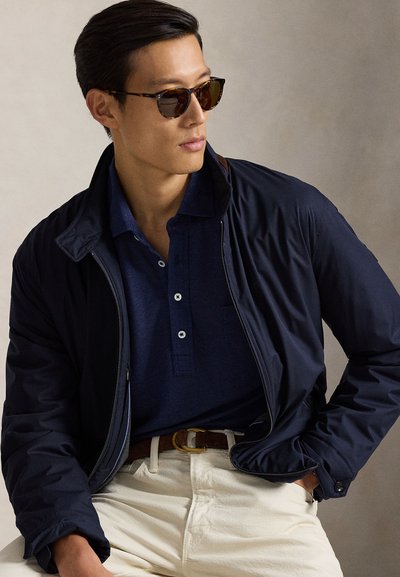 Ελαφρύ σακάκι ναυτικού πάνω από μια υφασμάτινη μπλε μπλούζα πόλο, συνδυασμένο με μπεζ παντελόνια και γυαλιά ηλίου σε σχέδιο χελώνας. Ορατή λεπτομέρεια με χρυσή αγκράφα ζώνης.