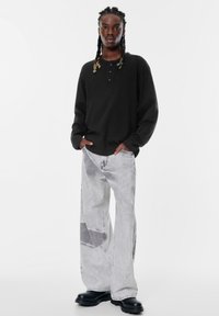 Schwarzes Langarm-Henley-Shirt mit drei Knöpfen, kombiniert mit hellgrauen, weit geschnittenen Jeans im Patchwork-Design und auffälligen schwarzen Schuhen.