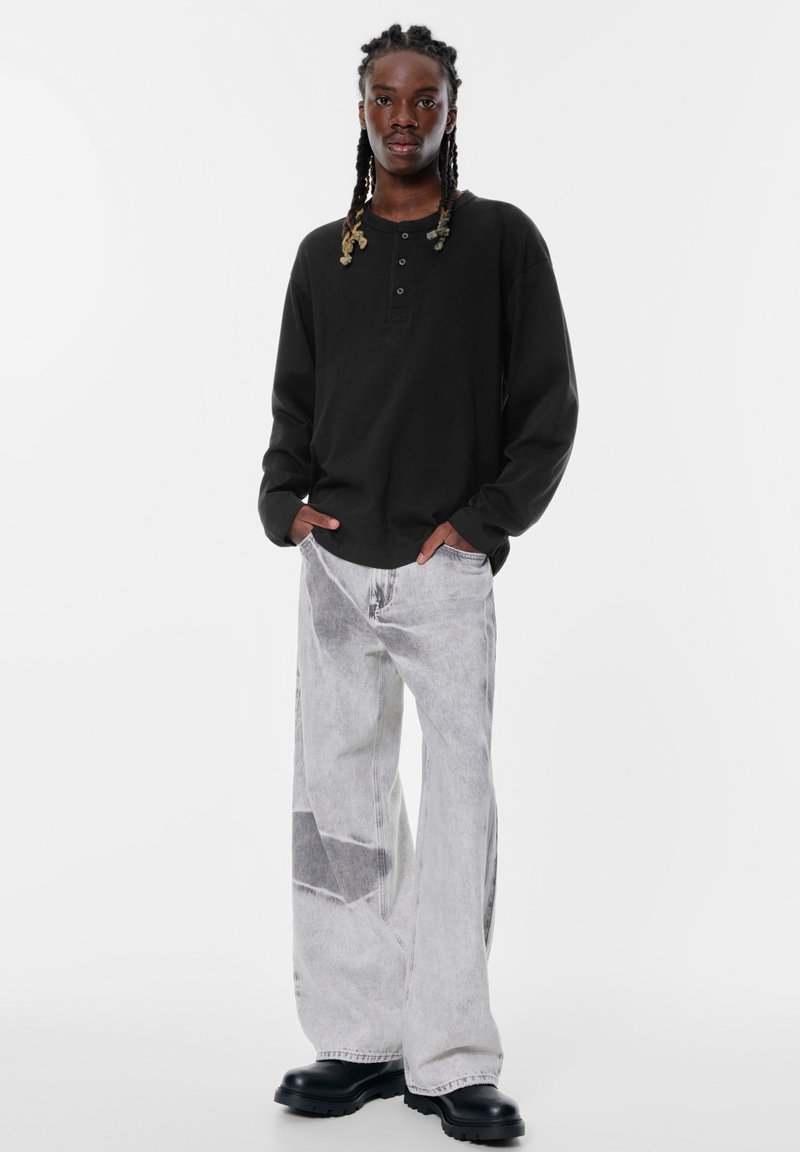 Schwarzes Langarm-Henley-Shirt mit drei Knöpfen, kombiniert mit hellgrauen, weit geschnittenen Jeans im Patchwork-Design und auffälligen schwarzen Schuhen.
