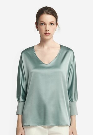 Satinblus i grönt med V-ringning, trekvartslånga ärmar och elastiska manschetter; slät textur och minimalistisk design.