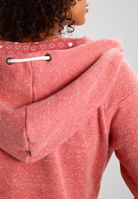 Sudadera con capucha de forro polar rojo de textura suave, con capucha con cordón, forro con patrón de corazones y ojales metálicos negros para el cordón.