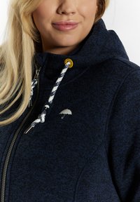 Jūrininkų gobtuvinė striukė, pagaminta iš tekstūruoto audinio. Puošiama sidabriniu skėčio logo, užtrauktuku, ir raštuotais raišteliais su geltonais akcentais.
