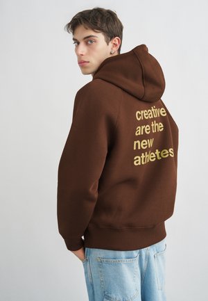 Fiatal férfi barna kapucnis pulóverben, amelyen az "A kreatívok az új sportolók" szöveg olvasható, és világoskék farmerben, váll fölött néz, egyszínű háttér előtt.