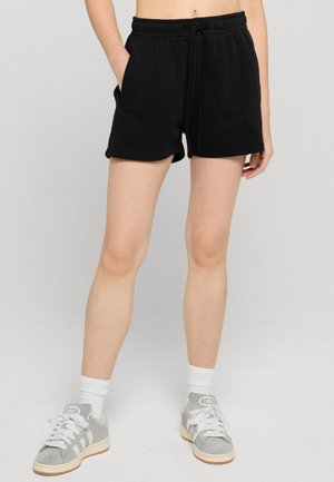 Personne portant un short noir à cordon, des chaussettes blanches et des baskets Adidas grises, debout avec une main dans la poche.