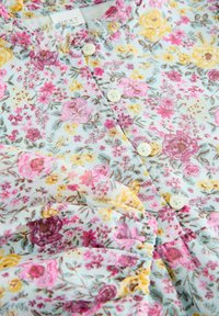 Blusa floreale con sfondo azzurro chiaro, caratterizzata da fiori rosa, gialli e marroni, bottoni bianchi e dettagli arricciati sulle maniche.