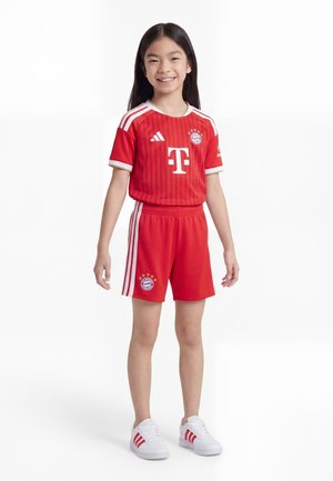 Uśmiechnięta dziewczyna stoi ubrana w czerwony strój piłkarski Bayernu Monachium z logo, białe spodenki w paski i białe trampki Adidas na jednolitym tle.