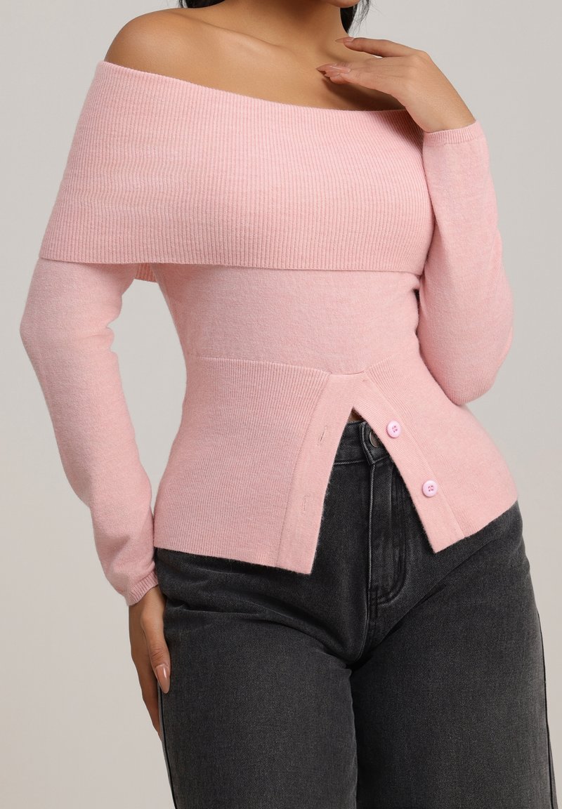 Femme portant un pull rose à col bateau avec des boutons à la taille, associé à un jean noir taille haute.