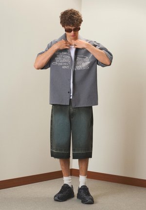 Ung mand iført solbriller, åben ternet skjorte med tekst, hvid t-shirt, lange jeansshorts, hvide sokker og sorte sneakers, der justerer kraven.