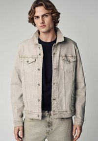 Lichtbeige denimjas met een klassieke kraag, twee borstzakken en knopen aan de voorkant. Draagt over een donker overhemd en heeft een zachte textuur.