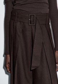 Bruine broek met een brede riem en een lang, gestrikt detail. De stof lijkt textuur en zacht, met plooien aan de voorkant voor extra detail.