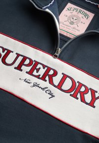 Sudadera de medio zipper en azul marino con rayas horizontales en blanco y rojo. Presenta "SUPERDRY" bordado en negrita y una etiqueta de logo sutil.