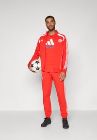 Rød treningsdrakt med hvite striper, med en logo på jakken og buksene. Kombinert med en rød T-skjorte og svarte joggesko. Holder en fotball.