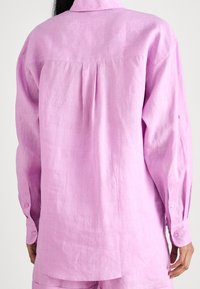 Light pink linen shirt with long sleeves, a folded collar, and buttoned cuffs. Features a pleat detail on the back and a curved hemline.