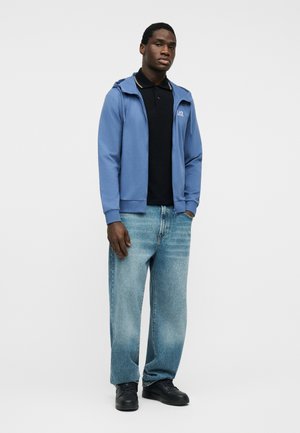 Hombre de pie vistiendo una sudadera azul con cremallera, polo negro, jeans holgados de mezclilla azul clara y zapatillas negras contra un fondo blanco liso.