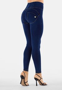 Korkeavyötäröiset, tummansiniset farkkuleggingsit, joissa on kaarevat takataskut. Sileä pinta ja ommellut yksityiskohdat, yhdistettyinä mustiin korkokenkiin.