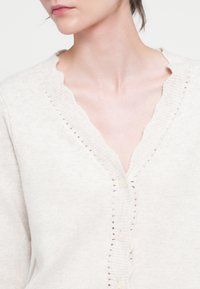 Jasno beżowy sweter zapinany na guziki z falistym dekoltem w kształcie litery V, z teksturowanymi, prążkowanymi brzegami oraz perłowymi guzikami. Zawiera małe wycięcia wzdłuż przodu.