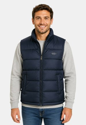 Homme souriant portant une veste matelassée bleu marine sur un pull gris clair et un jean bleu, debout devant un fond blanc uni.