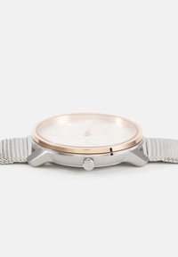 Montre ronde avec un bracelet en maille argentée, un cadran blanc, une lunette dorée et un boîtier argenté. Présente des indices d'heure minimalistes et une couronne.