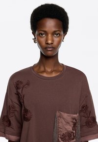 Bruine t-shirt met korte mouwen met bloemenborduursel aan de linkerkant en een satijnen zak met franjesdetails aan de voorkant.