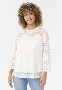Blusa bianca in pizzo con intricate fantasie floreali, bordi a ondulato e maniche a tre quarti, abbinata a jeans azzurri.