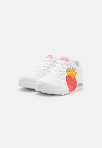 Zapatilla blanca con parte superior de malla y sintético, que presenta un vibrante diseño de corazones rojos y amarillos en el lateral, cordones planos y suela acolchada.