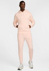 Felpa Nike rosa chiaro con tasca a marsupio frontale, pantaloni jogger rosa abbinati e sneakers grigie chiare. Presenta un logo Nike bianco sul petto.