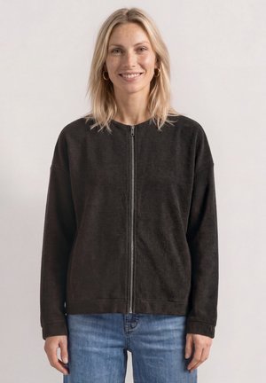 Blonde Frau lächelt, trägt goldene Creolen, eine schwarze Reißverschlussjacke und blaue Jeans, steht vor einem schlichten hellen Hintergrund.