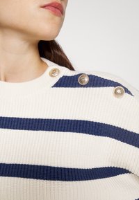 Cremfarbener Pullover mit navyblauen horizontalen Streifen, geribbelter Textur und dekorativen goldenen Knöpfen an der Schulter.