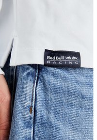 Balts kokvilnas t-krekls ar melnu etiķeti, uz kuras rakstīts "Red Bull Racing" baltā krāsā, savienots ar gaiši ziliem džinsiem.