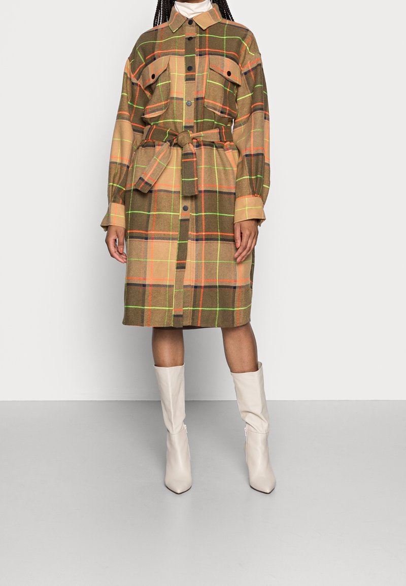 Koszula sukienka w kratę w brązowych, pomarańczowych i zielonych odcieniach, z paskiem w talii, kieszeniami na piersi i długimi rękawami. Połączona z białymi botkami.